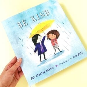Cover of book called Be Kind showing two girls on the cover, one is holding an umbrella over the other's head and it is raining