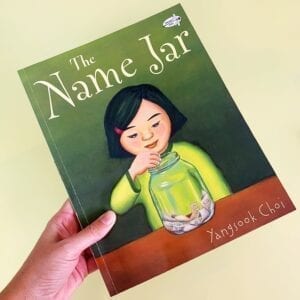 Cover of The Name Jar showing a girl putting paper into a jar on a table. The jar already has other small pieces of paper in it.