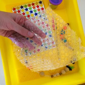 Hand holding bubble wrap with stickers, playdoh, and beads in a tub underneath the bubble wrap.