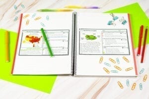 Notebook showing two exit ticket passages. One has an image of a frog and then other a map of the united states. The text is illegible because it is small. There are pencils and pens laying around the passages which are set atop an open notebook along with paper clips.