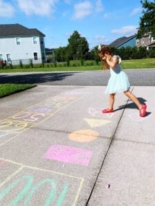 A little girl with a blue dress and brown shoes is preventing the summer slide by home learning on an obstacle course drawn from chalk
