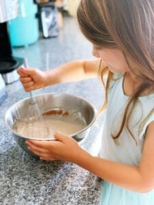 A little girl with brown hair and a blue dress is stirring cake mix with a whisk as she is helping her parents cook while learning math